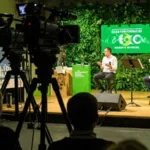 Photo d'un événement en présentiel, professionnel avec un bilan carbone réduit. Des caméras filment l'événement. Le décor est vert. L'écran sur scène et les kakémono présent sur place comportent un visuel éco responsable.
