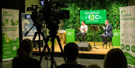 Photo d'un événement en présentiel, professionnel avec un bilan carbone réduit. Des caméras filment l'événement. Le décor est vert. L'écran sur scène et les kakémono présent sur place comportent un visuel éco responsable.